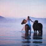 Eine junge Frau mit zwei Pferden im Wasser mit Bergkulisse