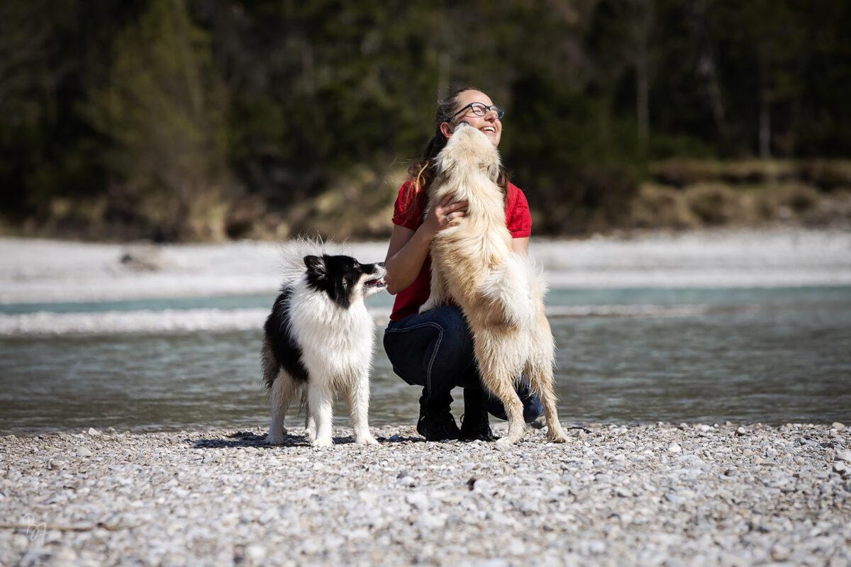 Foto einer Frau, die ihre beiden Hunde am Flussufer knuddelt