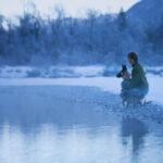 Eine Frau sitzt mit zwei Hunden an einem verschneiten Ufer