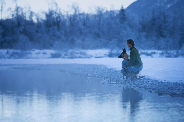 Eine Frau sitzt mit zwei Hunden an einem verschneiten Ufer
