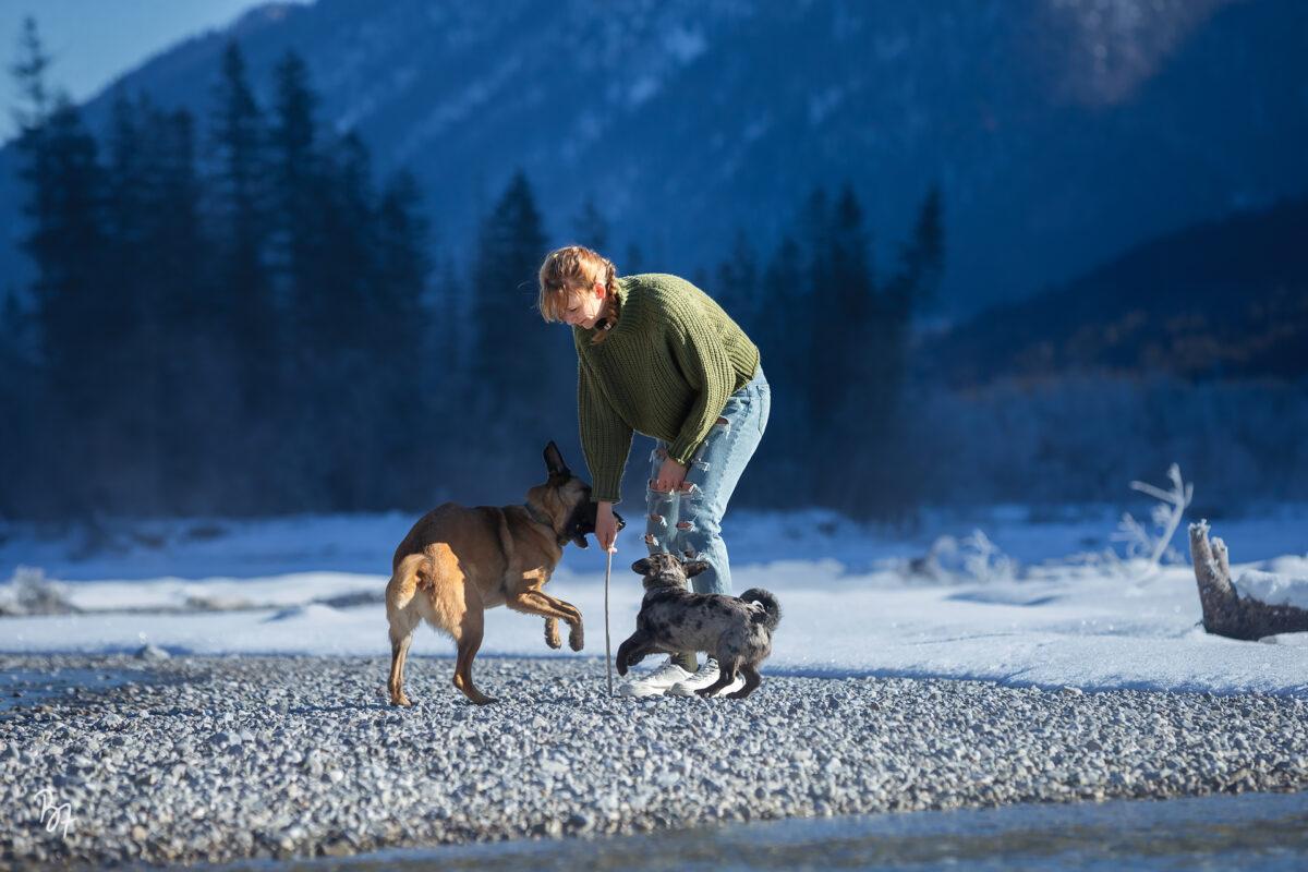 Eine Frau spielt mit ihren zwei Hunden an einem Ufer in winterlicher Berglandschaft